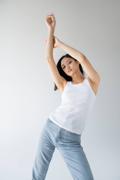 стройная азиатская женщина в белом топе и синих джинсах позирует с поднятыми руками, изолированными на сером - Фото, изображение