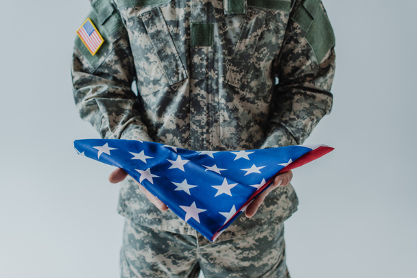 cropped view of American soldier in uniform holding folded flag of United States during memorial day isolated on grey  - Photo, Image