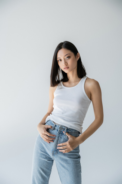 mladý a štíhlý asijský model pózující v modrých džínách a bílé tílko izolované na šedé - Fotografie, Obrázek