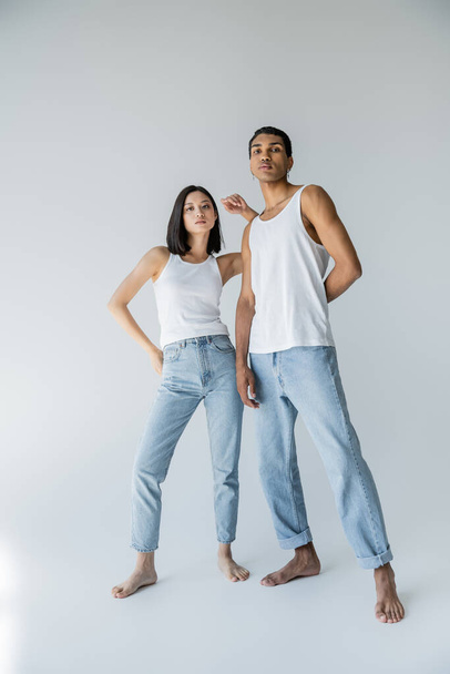 full length of barefoot interracial couple in blue jeans and white tank tops looking at camera on grey background - Fotó, kép