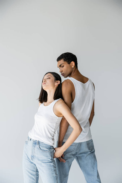 sensual asian woman with closed eyes leaning on african american man in white tank top and jeans isolated on grey - Photo, Image