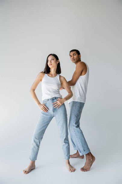 full length of stylish asian woman posing with thumbs in pockets of jeans near barefoot african american man on grey background - Photo, Image