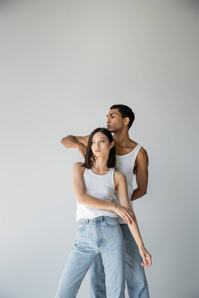 young african american guy touching neck of asian woman in white tank top and jeans isolated on grey - Фото, изображение