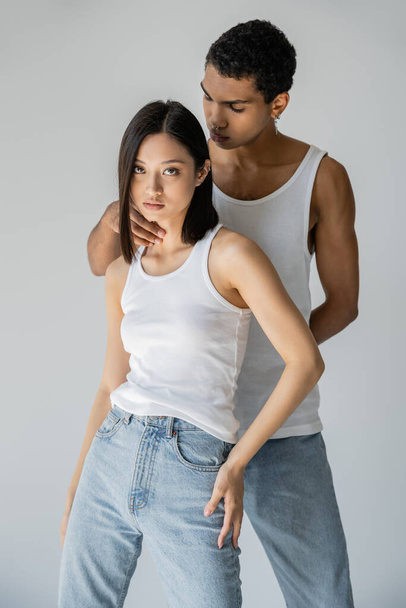 young african american man touching neck of asian woman in white tank top and jeans isolated on grey - Фото, изображение
