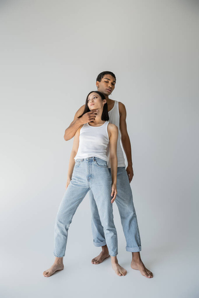 full length of stylish african american man touching chin of barefoot asian woman in tank top and jeans on grey background - Fotografie, Obrázek