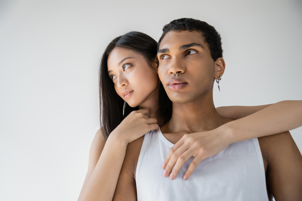 sensual asian woman embracing stylish african american man in white tank top while looking away isolated on grey - Фото, изображение