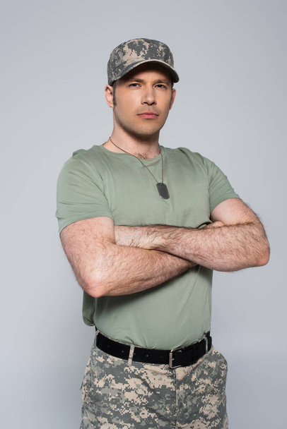 στρατιώτης με t-shirt και καπέλο στέκεται με διπλωμένα χέρια κατά τη διάρκεια της ημέρας μνήμης απομονωμένος σε γκρι  - Φωτογραφία, εικόνα