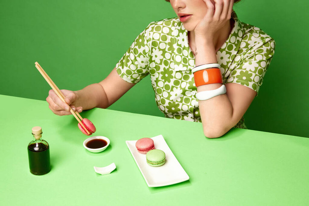 Cropped image of girl putting sweet macarons into soy sauce with chopsticks over green studio background. Food pop art photography. Concept of retro style, creative vision, imagination. Sweet and salt - Foto, afbeelding