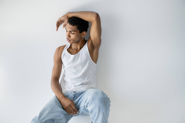 pensive african american man in white tank top and blue jeans sitting on grey background - Фото, зображення