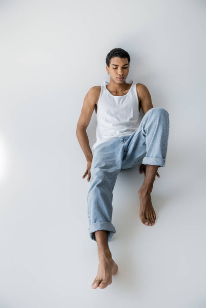  high angle view of young and barefoot african american man posing in tank top and jeans on grey background - Photo, Image