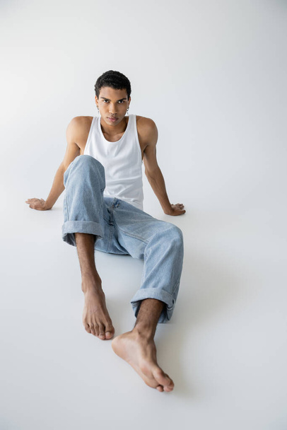 full length of stylish african american man in blue jeans and tank top sitting and looking at camera on grey background - Fotó, kép