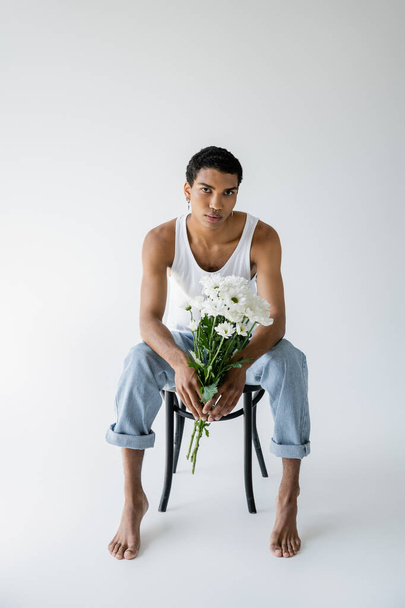 full length of african american man in tank top and jeans holding flowers and looking at camera while sitting on grey background - Photo, Image