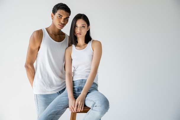 young asian woman in jeans and white tank top sitting on chair near young african american guy isolated on grey - Fotó, kép