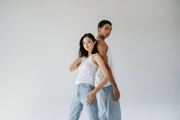 young and stylish interracial couple in blue jeans and white tank tops looking at camera isolated on grey - Fotografie, Obrázek