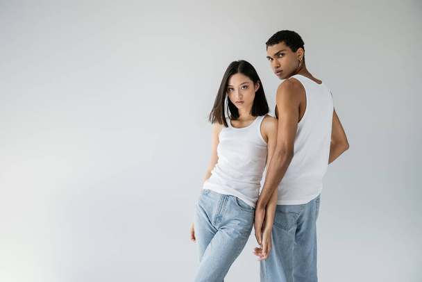 brunette asian woman and african american guy in blue jeans and white tank tops looking at camera isolated on grey - Fotografie, Obrázek