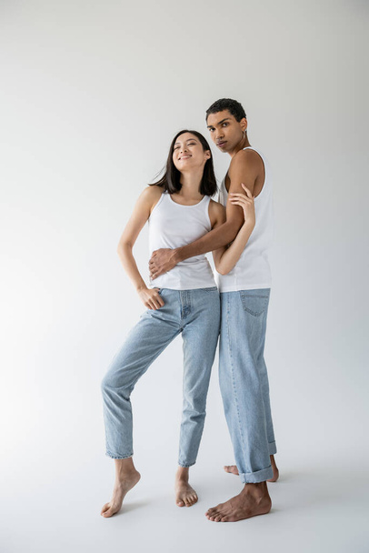 full length of african american man in tank top and jeans embracing cheerful asian model on grey background - Φωτογραφία, εικόνα
