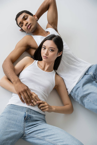 top view of young asian woman in tank top and jeans lying near african american man and looking at camera on grey background - 写真・画像