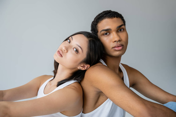 brunette asian woman and stylish african american man with silver piercing sitting back to back isolated on grey - Foto, Imagem