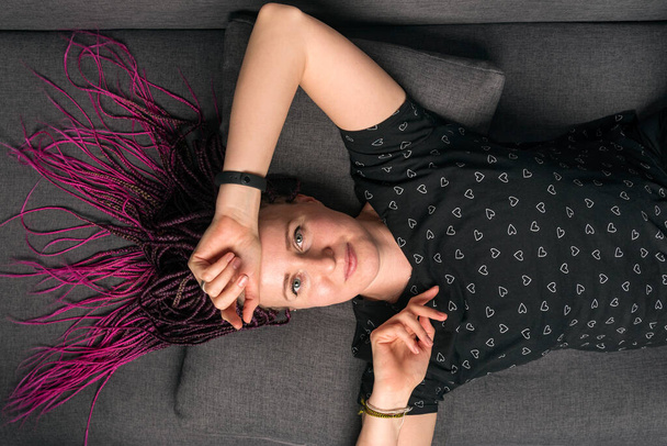 Portrait of pritty casually dressed caucasian young woman with dreadlocks looking into camera and lying on the sofa - Photo, Image