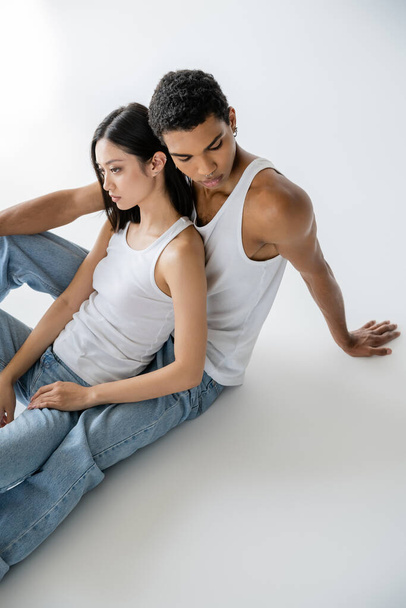 vista ad alto angolo di giovane coppia interrazziale in canotte bianche e jeans seduti su sfondo grigio - Foto, immagini
