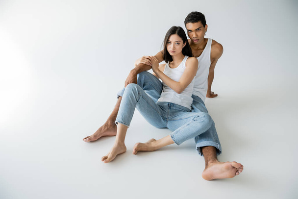 full length of barefoot multiethnic couple sitting in blue jeans and white tank tops on grey background - Photo, Image