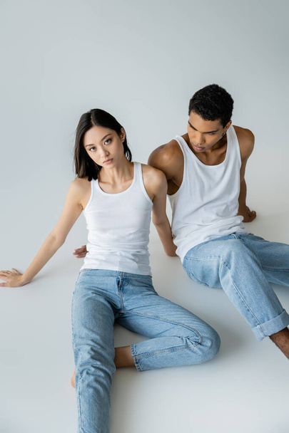 slim asian woman in blue jeans and white tank top looking at camera near african american guy on grey background - Foto, Imagem