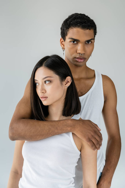 afro americano cara no branco tanque top abraçando morena asiático modelo e olhando para câmera isolado no cinza - Foto, Imagem