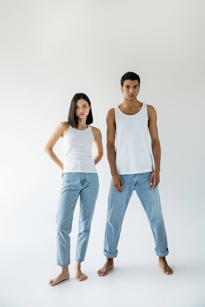 full length of young and barefoot multiethnic couple posing in white tank tops and blue jeans on grey background - Fotografie, Obrázek