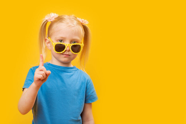 Portrait of little girl wears blue T-shirt and yellow sunglasses on bright yellow background. Child raised her index finger. Copy space, mock up. - Photo, Image