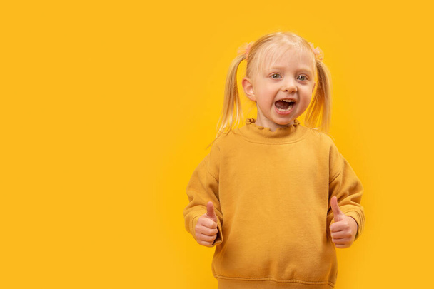 Funny blonde girl in yellow sweatshirt shows two fingers up like gesture. Yellow background. Copy space, mock up. - Photo, Image