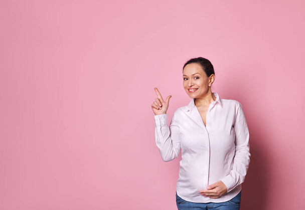 Πολυεθνική ευτυχισμένη έγκυος γυναίκα κρατώντας το χέρι στην κοιλιά της, χαμογελώντας ένα όμορφο χαμόγελο toothy κοιτάζοντας κάμερα, δείχνοντας δείκτη της σε αντίγραφο διαφημιστικό χώρο σε ροζ φόντο. Άδεια μητρότητας. Κύηση - Φωτογραφία, εικόνα