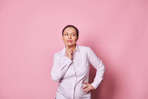 Perplexed doubtful multi ethnic gravid woman holding finger near her lips, thoughtfully looking away at copy space, reasoning, on pink isolated backdrop. Emotional portrait of pensive pregnant female - 写真・画像