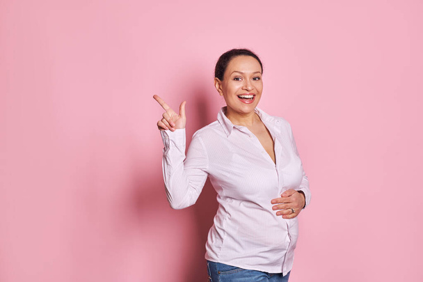Mulher grávida feliz tocando suavemente sua barriga e sorrindo olhando para a câmera, apontando seu dedo indicador para espaço publicitário cópia em fundo rosa isolado. Gravidez saudável e conceito de maternidade - Foto, Imagem