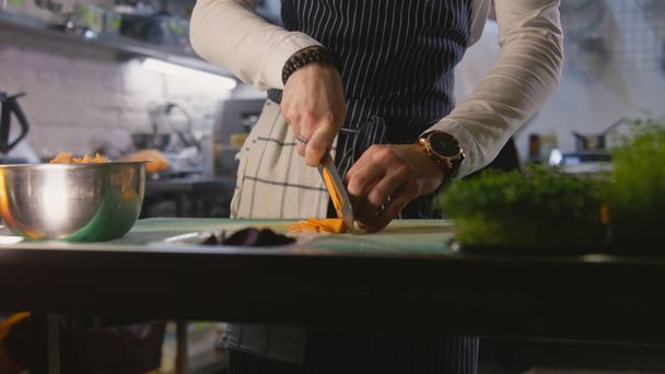 Hands close-up of professional male chef standing by kitchen table and cutting sweet potato. Busy coworker prepares dishes at background. Concept of teamworking in restaurant. Slow motion. Dolly zoom. - Photo, Image