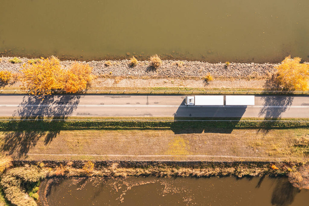 El camión sale a la carretera en otoño. transporte de coches. Camión con semirremolque en color gris. Camión de transporte conduce en otoño junto al lago en un día soleado. - Foto, imagen
