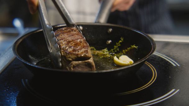 The chef cooks delicious meal on stove. The cook fries piece of juicy pork meat on frying pan with rosemary, pours oil using spoon. Process of steak cooking. Restaurant with professional kitchen. - Фото, изображение
