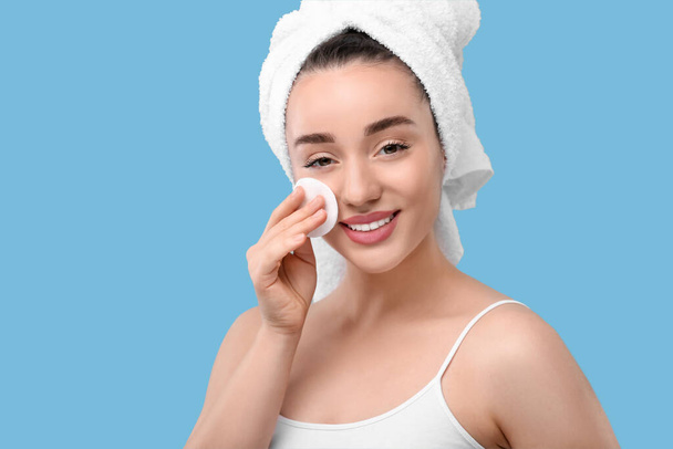 Beautiful woman in terry towel removing makeup with cotton pad on light blue background - Фото, изображение