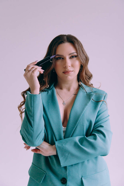 Luxurious girl with beautiful makeup and hairstyle, permanent makeup specialist, holds tools in her hands. Photoshoot in a photo studio in a suit, jeans and a shirt. - Foto, afbeelding