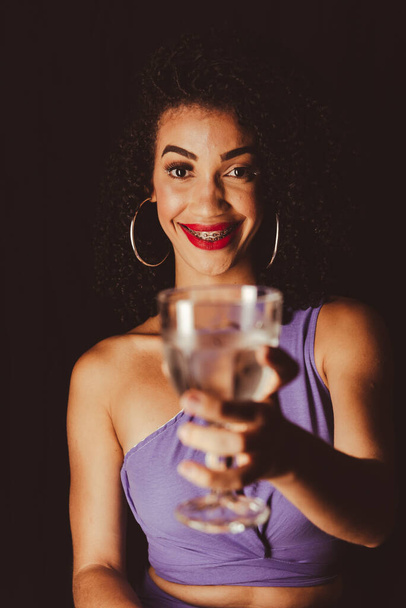 Молодая красивая улыбающаяся женщина показывает стеклянную чашку. Изолированный чёрный фон. - Фото, изображение