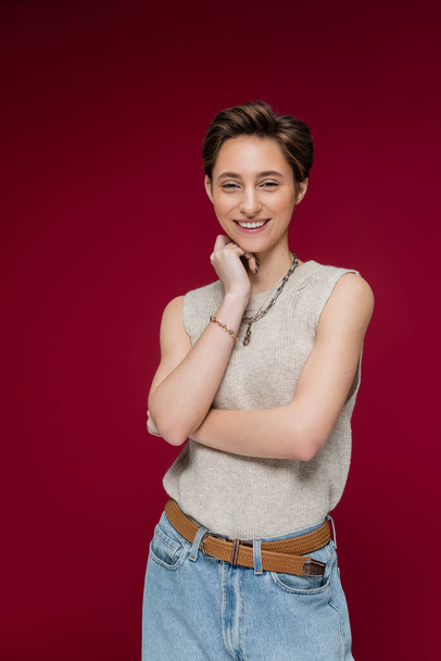 cheerful young woman with short hair smiling and looking at camera on dark red background - Photo, Image