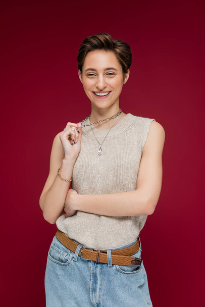 pretty young woman with short hair smiling and pulling necklace chain on dark red background - Фото, зображення