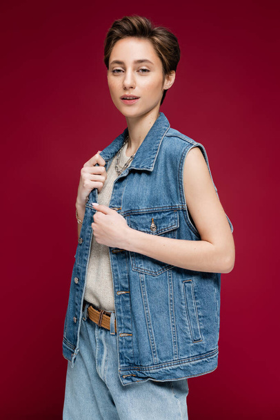 stylish young woman with short hair posing in denim vest on dark red background - Photo, Image
