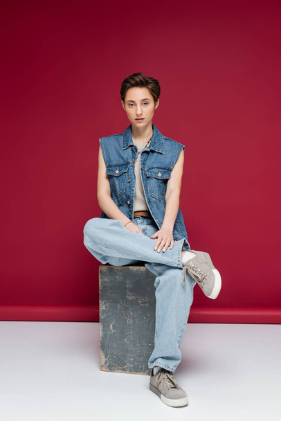 full length of stylish model with short hair sitting in denim outfit on dark red background - 写真・画像