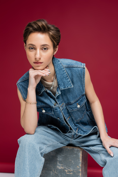 stylish young model with short hair sitting in denim outfit on dark red background - Foto, afbeelding