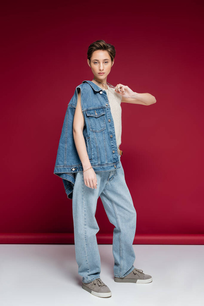 full length of pretty young woman in denim vest pulling necklace chain while posing on dark red background - Фото, изображение