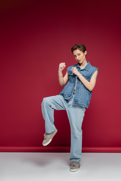 full length of sportive woman with short hair standing with clenched fists while fighting on dark red background  - Photo, Image