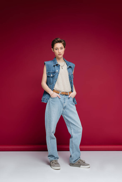 full length of young model in denim outfit standing with hands in pockets on dark red background - Foto, Imagem