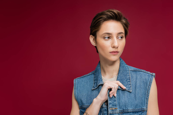 stylish model in denim outfit with vest looking away on burgundy background  - Фото, зображення