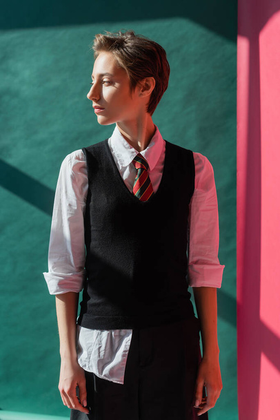 stylish student with short hair standing in school uniform on pink and green  - Фото, зображення