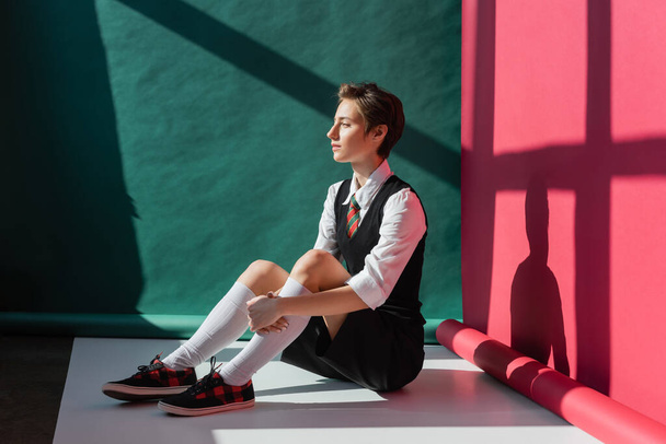 full length of stylish young woman with short hair sitting in school uniform on green and pink background  - Photo, Image